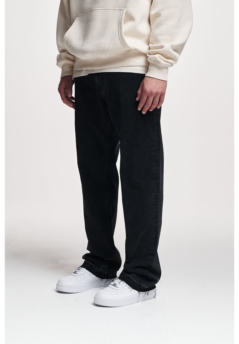 Personne portant des baskets blanches, un jean noir ample et un sweat à capuche beige, debout sur un fond clair uni.