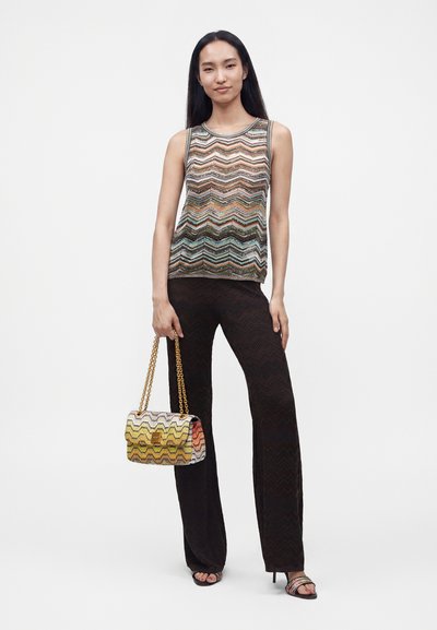 Femme debout portant un top sans manches à motif zigzag, pantalon noir à motif zigzag, tenant un sac à main multicolore à motif avec des anses en chaîne.