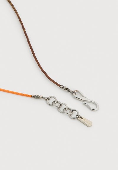 Δύο άκρα περιδέραιου με χάντρες, το ένα καφέ με κούμπωμα γάντζου από ασήμι, το άλλο πορτοκαλί με ασημένιες αλυσίδες και μικρή ετικέτα με το σήμα της μάρκας.