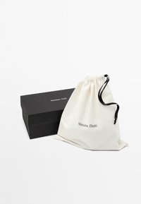 Sac en coton avec cordon de serrage en crème portant "Massimo Dutti" imprimé en noir, à côté d'une boîte rectangulaire noire mate avec des bordures dorées.