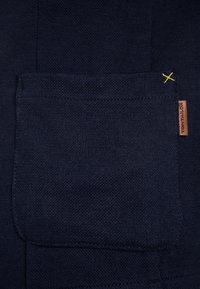 Tasca in tessuto blu navy con superficie testurizzata, caratterizzata da un dettaglio di cucitura gialla e un piccolo cartellino in pelle etichettato "TOM TAILOR."