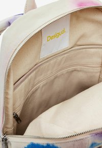 Canvas-Rucksackinnenraum in Creme mit einem bunten Batikmuster. Verfügt über eine Reißverschlusstasche und ein beschriftetes Etikett. Weiche Textur.