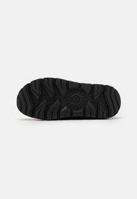 Black rubber shoe sole with a rugged tread pattern, featuring deep grooves and a circular emblem at the center for traction and grip.
