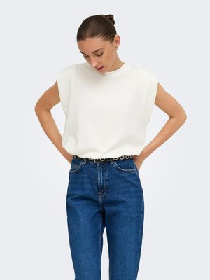 Kvinde med håret sat op i en knold iført en hvid ærmeløs top med polstrede skuldre, stoppet ned i højtaljede blå jeans med sort bælte.