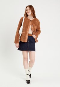 Veste en polaire marron avec fermeture à boutons, associée à un col roulé beige et une jupe en denim noir. Les accessoires comprennent un sac à épaule blanc et des chaussettes crème.