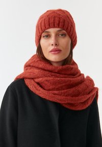 Cappello lavorato a maglia di colore rosso-arancio con design a costine e sciarpa morbida e texturizzata coordinata, drappeggiata attorno al collo, abbinata a un cappotto nero.