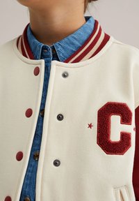 Krämfärgad varsityjacka med randig ribbad krage i rött och vitt, tryckknappar, ett rött filté brev "C" och en jeansskjorta lagd under.