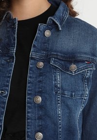 Tummansininen farkkutakki, jossa on hopeiset yksityiskohdat. Varustettu rintataskuilla, nappikiinnityksellä ja perinteisellä kauluksella. Kankaan pinta on teksturoitu.