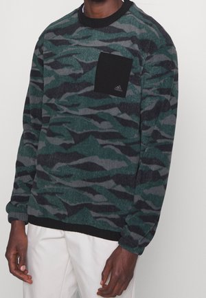 Mann trägt einen grünen und schwarzen Pullover mit Camouflage-Muster, langen Ärmeln, schwarzer Brusttasche und weißen Hosen.