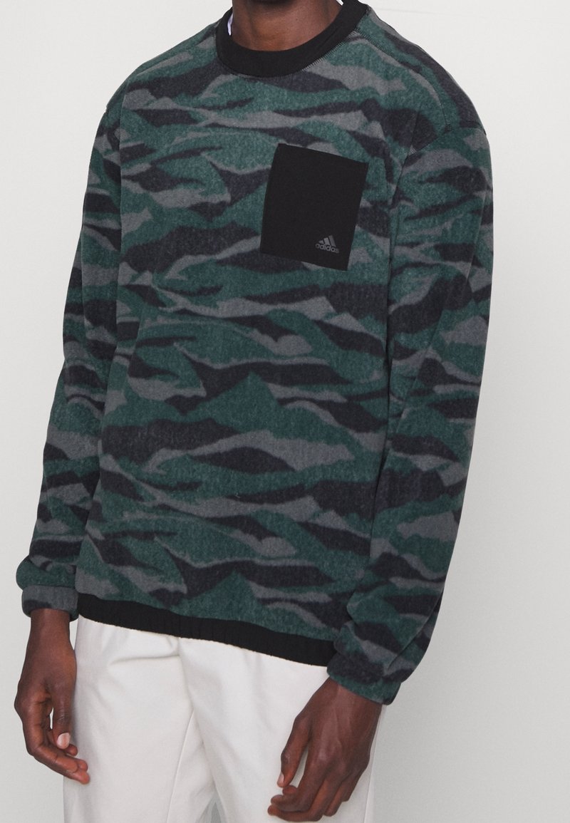 Homme portant un pull à manches longues vert et noir à motif camouflage avec une poche noire sur la poitrine et un pantalon blanc.