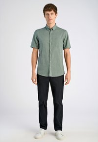 Camisa de botones de manga corta color verde claro con un bolsillo en el pecho, combinada con pantalones negros y zapatillas blancas. Textura suave, diseño casual.