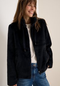 Veste zippée en fausse fourrure noire avec un col montant, dotée de cordons de serrage et de deux poches latérales, associée à un léger pull et à un jean.