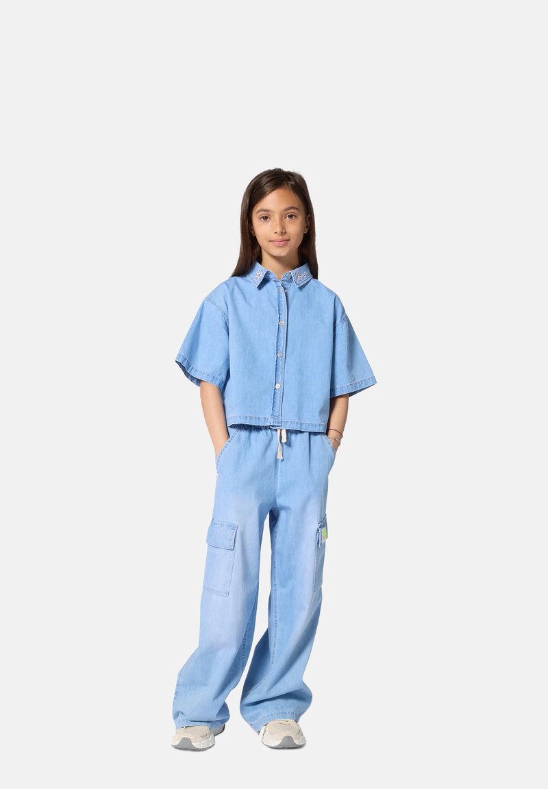 Chemise courte en denim bleu clair avec manches courtes et fermeture à boutons sur le devant, associée à un pantalon cargo bleu clair ample et des baskets.