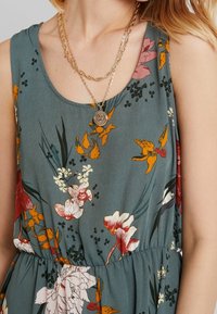 Květinové šaty z teal látky s barevnými květy v červené, žluté a bílé barvě. Vybavené náhrdelníkem s kulatým přívěskem a úzkým pasem.