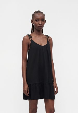 Femme mince avec des cheveux tressés portant une robe noire sans manches ample avec des bretelles nouées et un ourlet à volants, debout devant un fond clair uni.