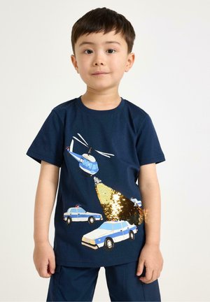 Ung dreng iført en mørkeblå skjorte med politibiler og en helikoptergrafik, hvor ét billys er lavet af skinnende gyldne palietter.