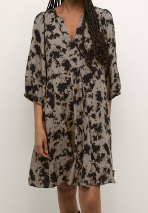 Kvinde iført løs knælængde kjole med sort og gråt abstrakt tie-dye mønster og trekvartlange ærmer, stående mod en ensfarvet baggrund.