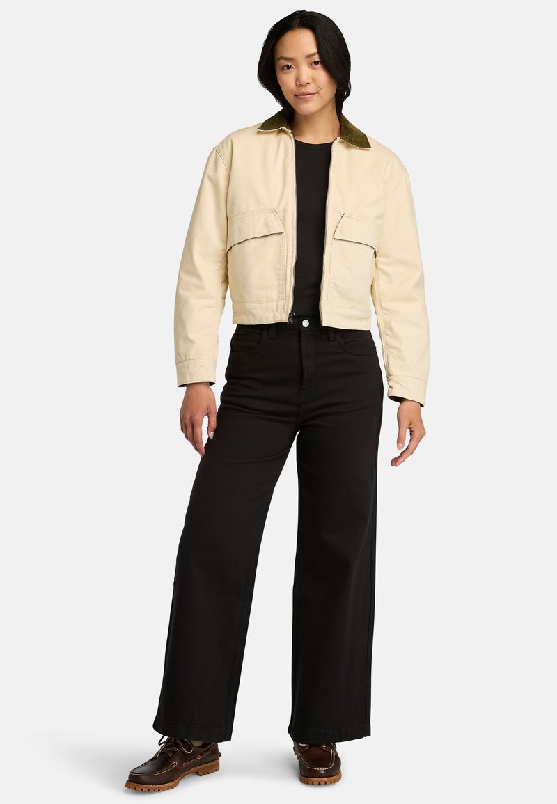 Giacca beige accorciata con colletto in cotone verde, due tasche frontali, indossata sopra una camicia nera, abbinata a pantaloni neri a gamba larga e stivali.