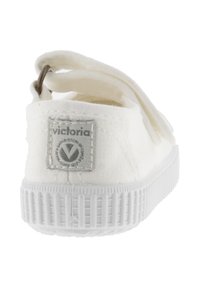 Zapatilla de lona blanca con suela texturizada y parche de logo gris en la parte trasera; cuenta con un lazo de tela y un diseño sencillo de fácil puesta.