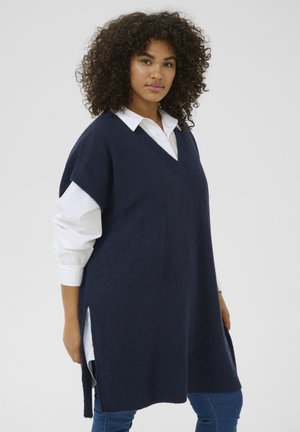 Granatowy sweter z dzianiny z dekoltem w szpic, z bocznymi rozcięciami i oversize'owym krojem, noszony na białej koszuli z kołnierzykiem. Miękka tekstura i swobodny fason.