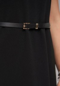 Schwarzer Ledergürtel mit einer metallischen goldenen Schnalle, verstellbares Design mit mehreren Löchern, befestigt an einem glatten Stoffkleidungsstück.
