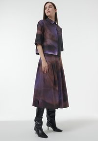 Camisa corta de estampado púrpura y marrón con cuello, combinada con una falda midi plisada. La modelo lleva botas de tacón negras hasta la rodilla.