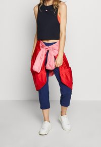 Černý crop top, volná červená a růžová bunda uvázaná v pase, modré zkrácené kalhoty a bílé tenisky. Materiál je prodyšný a barevný.