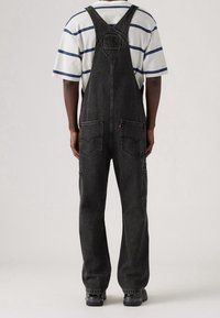 Svarta denimhängselbyxor med justerbara remmar, fram- och bakfickor, samt en lös passform. Kombinerat med en vit randig skjorta under.