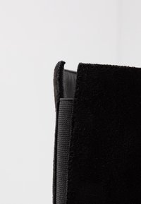 Botín negro de ante con una superficie texturizada, que presenta un panel de tela lisa en el costado y un diseño estructurado y minimalista.