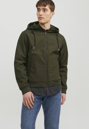 Jeune homme portant une veste zippée à capuche vert olive par-dessus une chemise à carreaux et un jean bleu, regardant sur le côté avec un fond uni.