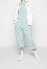 Mintově zelený teplákový komplet s bílým logem, obsahující zkrácený top a kalhoty s elastickým pasem. Doplněno bílou lehkou bundou a teniskami.