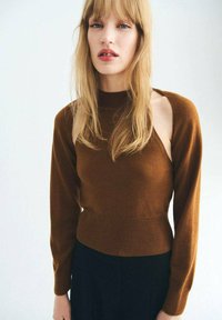 Junge Frau mit langen blonden Haaren, die einen braunen Pullover mit ausgeschnittenen Schultern und schwarze Hosen trägt, steht vor einem schlichten hellen Hintergrund.