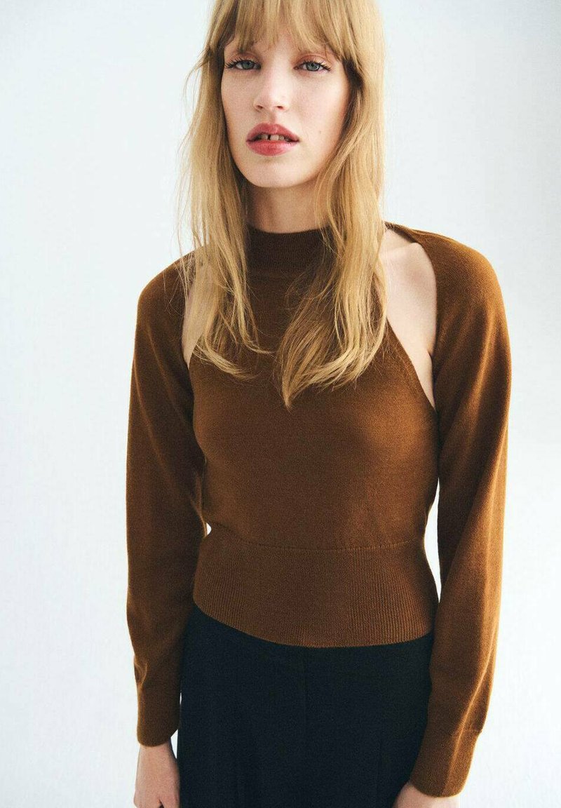 Junge Frau mit langen blonden Haaren, die einen braunen Pullover mit ausgeschnittenen Schultern und schwarze Hosen trägt, steht vor einem schlichten hellen Hintergrund.