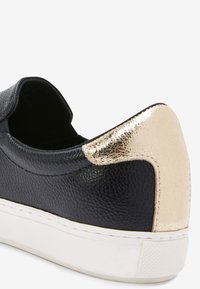 Černá kožená sneaker s texturovaným povrchem, s akcentem na zlatém kovovém podpatku a bílou gumovou podrážkou.