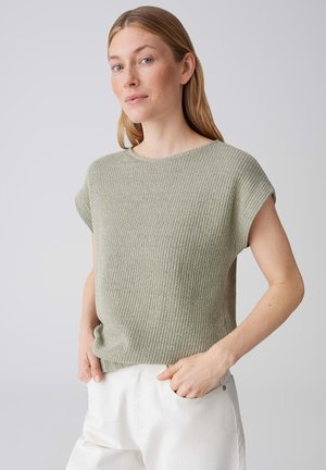 Femme aux longs cheveux blonds portant un haut en tricot sans manches vert clair et un pantalon blanc, les mains dans les poches, sur un fond uni.