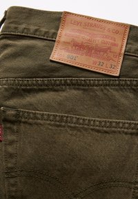 Poche arrière du jean Levi's 501 vert olive avec une étiquette en cuir marron affichant la marque, la taille W32 L32, et le logo signature des deux chevaux.