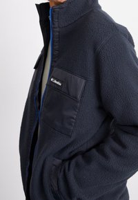 Granatowa polarowa kurtka z wysokim kołnierzem, zamkiem błyskawicznym z przodu i teksturowanym wykończeniem. Posiada gładką kieszeń na piersi z logo.