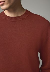 Röd stickad tröja med rund ribbad halsringning och strukturerad tyg. Visar en enkel design med subtilt synliga sömdetaljer.