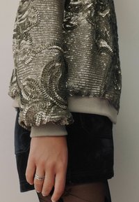 Silver sequin pullover with floral texture and ribbed cuffs, paired with a black velvet skirt; hand adorned with a silver ring.