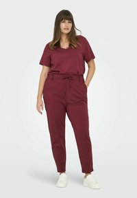 Tummanpunainen asu, jossa on lyhythihainen V-kaula-aukkoinen t-paita ja kiristysnyörillä varustetut housut. Sileä kangas, rento istuvuus ja valkoiset tennarit täydentävät kokonaisuuden.