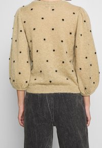 Pull en maille beige à pois noirs ; présente un col rond, des manches bouffantes et un ourlet côtelé. Tissu doux et texturé.