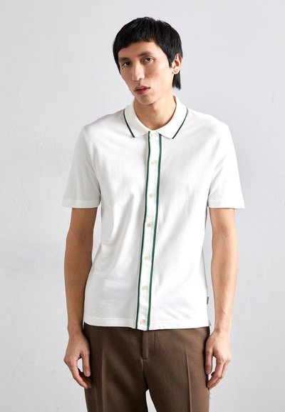 Polo shirt blanc à manches courtes avec piping vert, patte de boutonnage sur le devant et texture légère, associé à un pantalon marron ajusté.
