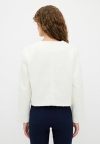 Suéter blanco corto de tela texturizada, con mangas largas y dobladillo recto. Combinado con pantalones ajustados azul marino, mostrando vista trasera.
