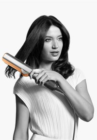 Haarstyler mit einem silbernen Gehäuse und orangen Akzenten, ausgestattet mit einer strukturierten Grifffläche und einem langen Kabel, gehalten von einer Person mit gestyltem Haar.