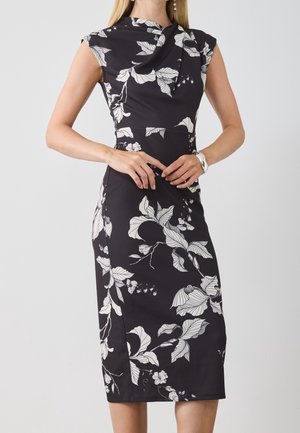 Czarna sukienka o długości do kolan z białym kwiatowym wzorem, z krótkimi rękawami typu cap i drapowanym dekoltem, noszona przez kobietę z splecionymi rękami.