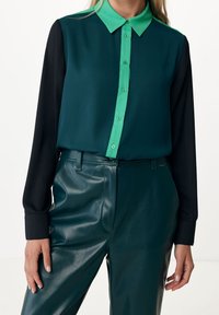 Dark green and black blouse with a green collar, button-down front, and long sleeves. Paired with glossy green leather pants.