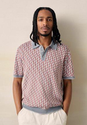 Man with shoulder-length dreadlocks wears a short-sleeve geometric-patterned polo shirt and light-colored pants, hands in pockets.