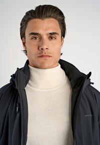 Svart jacka med hög krage, strukturerad ljus cream polotröja under. Enkelt design, slätt tyg, minimala metalldetaljer.