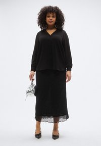 Μαύρο πουκάμισο με κουμπιά και μακριά μανίκια, συνδυασμένο με μια μαύρη, λαμπερή μίντι φούστα που έχει διάφανο κάτω στρώμα. Το μοντέλο κρατάει μια ασημί τσάντα χειρός.