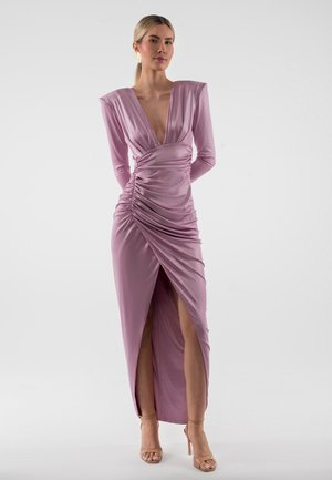 Žena oblečená v dlhý rukáv, nariasené levanduľové šaty s hlbokým výstrihom do V a rozparkom nad stehnom, stojaca pred jednoduchým svetlosivým pozadím.